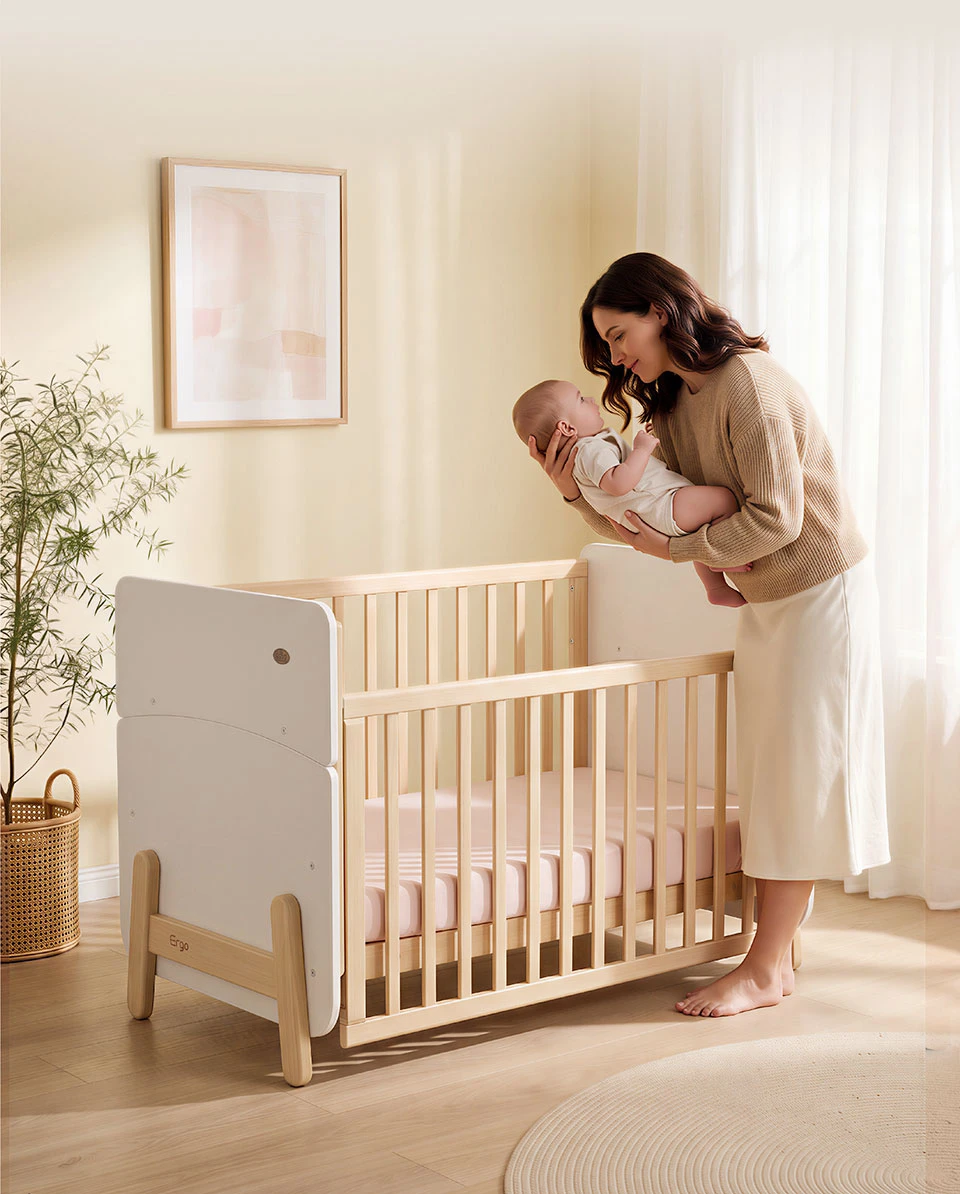 Mother lifting a baby above a wooden cot with white end panels and natural timber slatted sides in a sunlit nursery Mother lifting a baby above a wooden cot with white end panels and natural timber slatted sides in a sunlit nursery