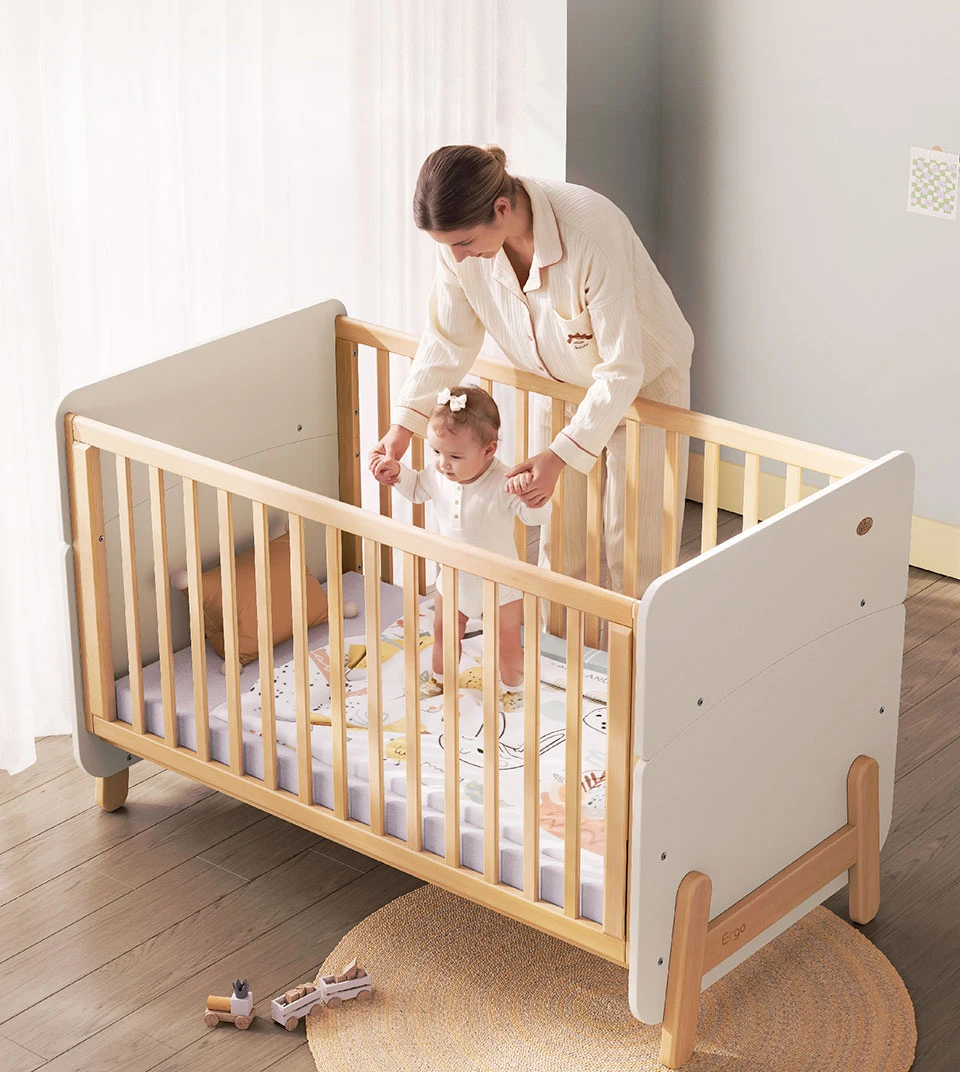 Mother holding the hands of a baby standing inside a wooden cot with slatted sides and white end panels Mother holding the hands of a baby standing inside a wooden cot with slatted sides and white end panels